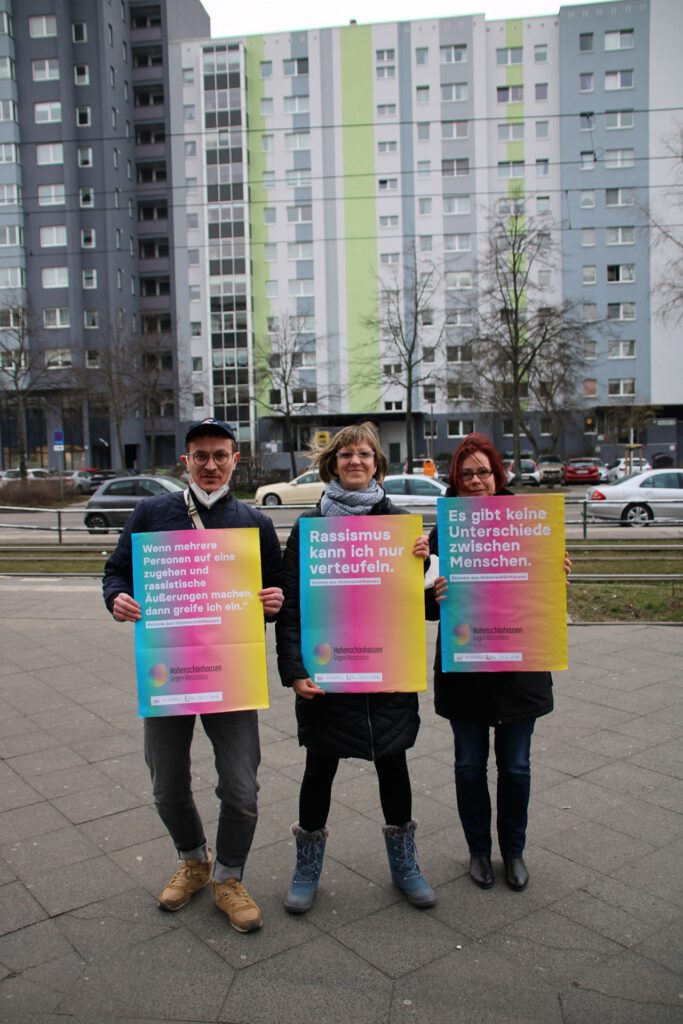 3 Personen auf der Straße halten bunte Plakate mit Sprüchen gegen Rassismus