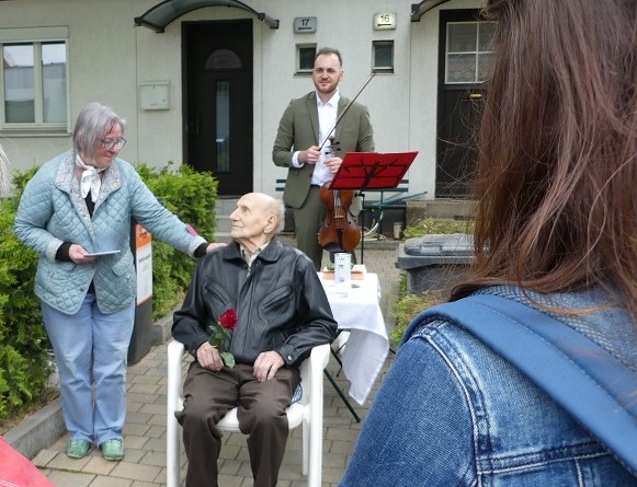Ein Alter Mann sitzt auf einem Stuhl mit einer roten Rose in der Hand, Dagmar Potzsch und weitere Teilnehmende stehen in der Nähe