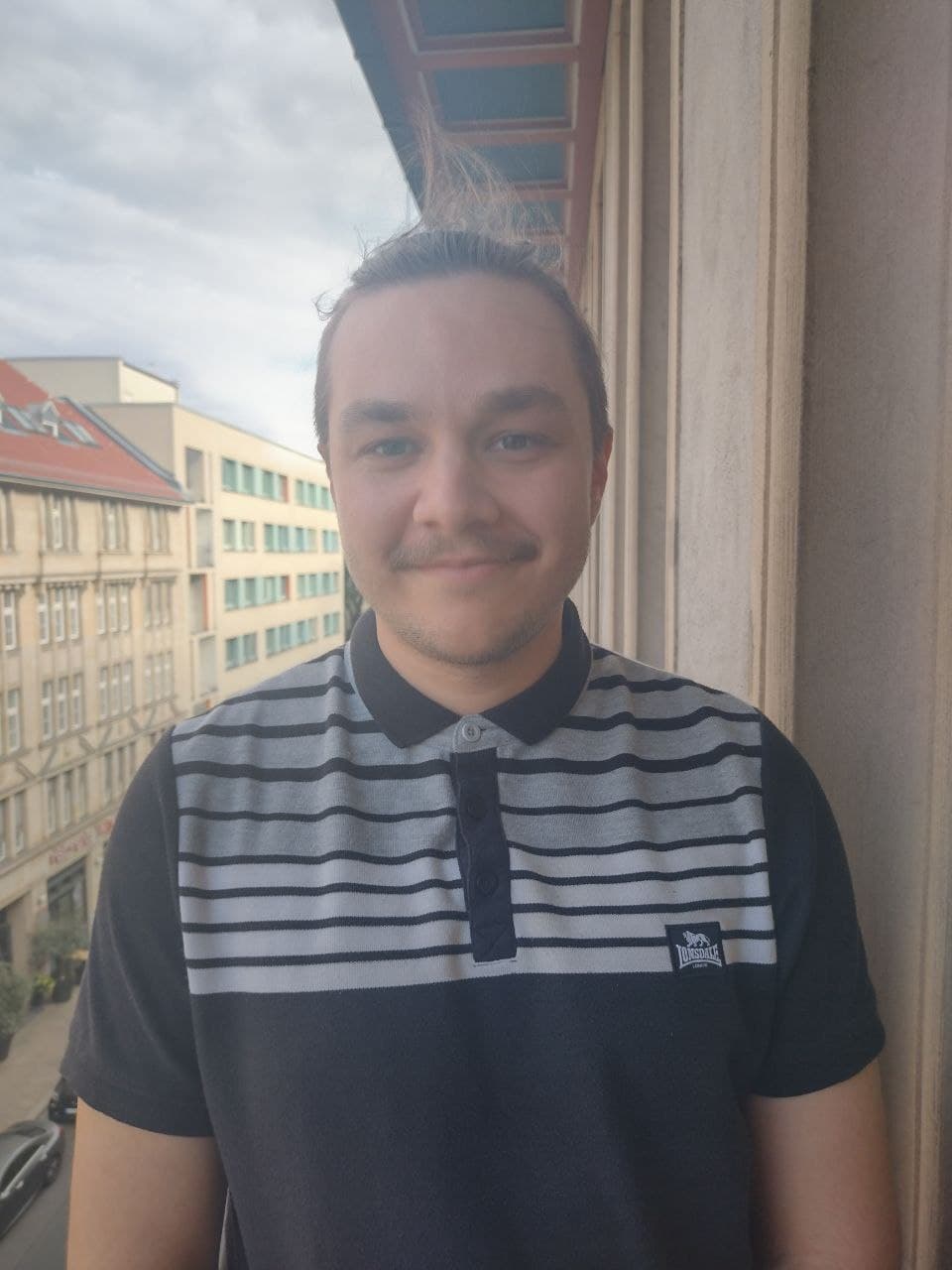 Portrait eines jungen Mannes mit kurzen dunklen Haaren und Schnurrbart mit Häusern im Hintergrund