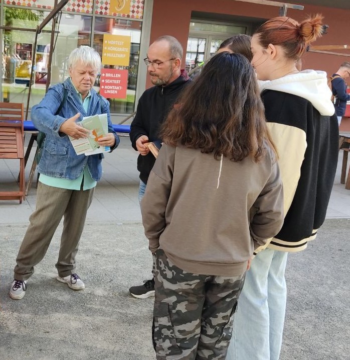 Eine Frau mit kurzen weißen Haaren undterhält sich mit jungen Menschen und zeigt einen Flyer