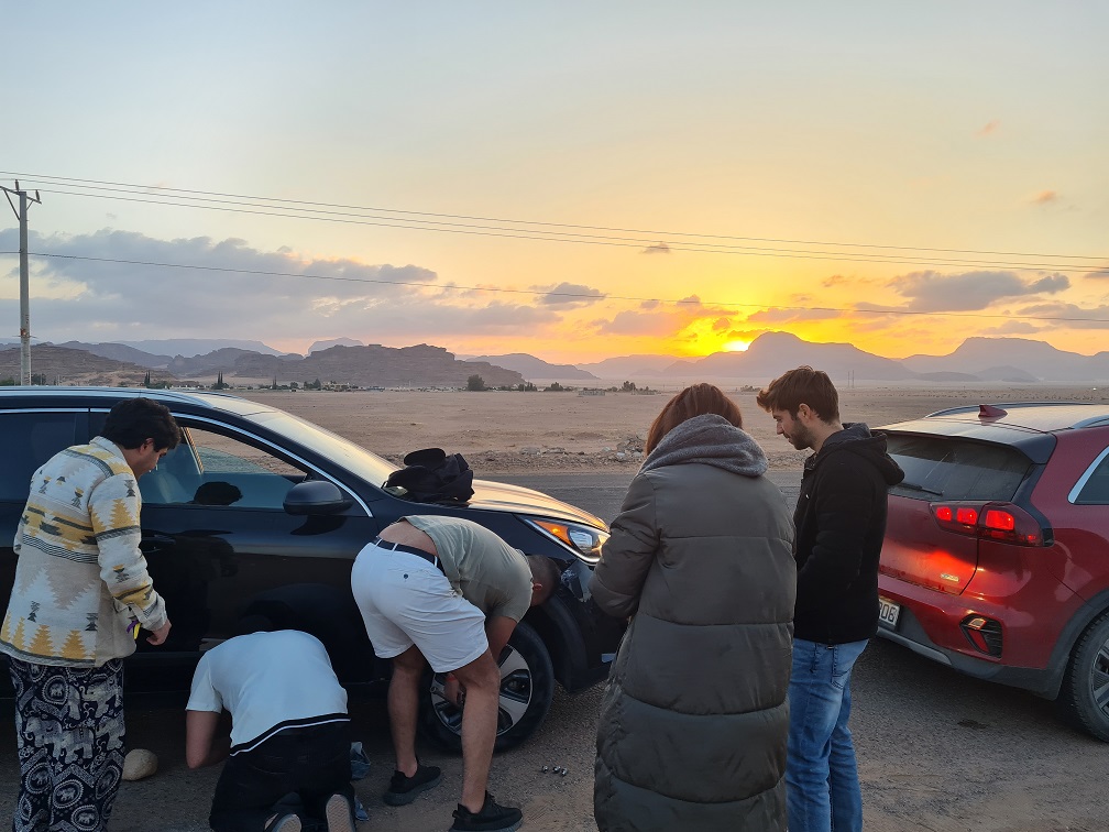 Menschen stehen gebückt vor einem Auto in der Wüste und arbeiten am Reifen