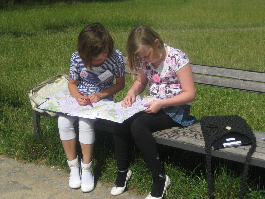 Zwei Kinder sitzen im Grünen auf einer Bank und schauen in eine große Karte, 