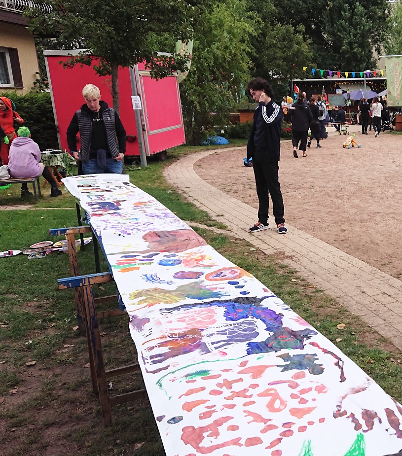 Eine lange bemalte Papierrolle liegt auf langen Tischen, Menschen stehen davor