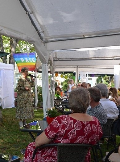 Eine Frau im Sommerkleid steht vor einer Gruppe sitzender Menschen unter einem Zeltdach im Sommer