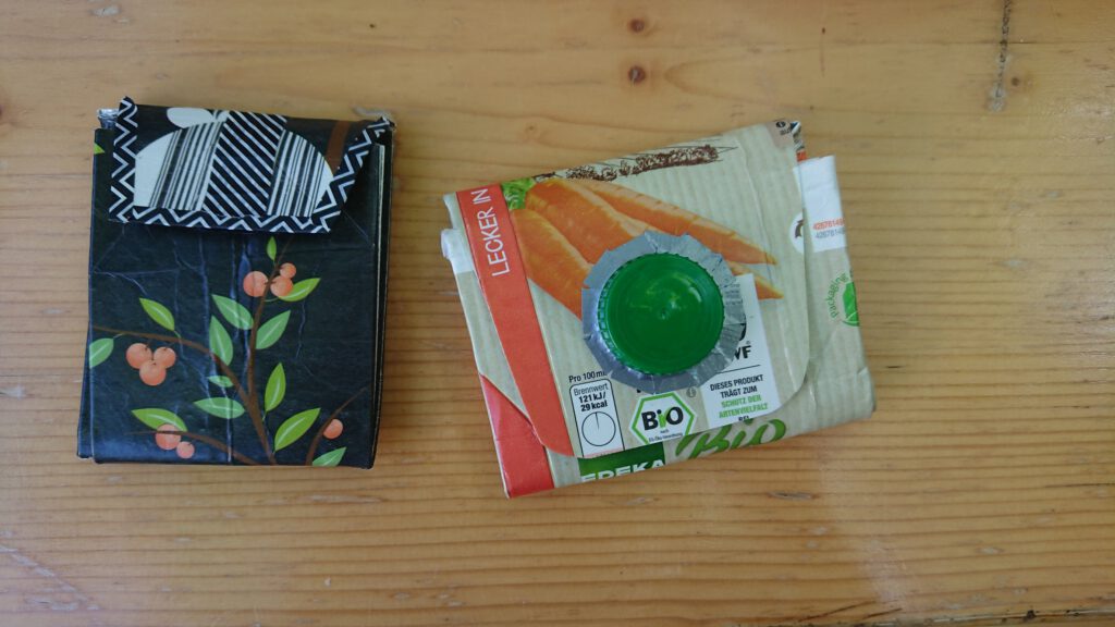 Zwei Geldbeutel aus Tetra Packs. Links ein schwarzer Geldbeutel, etwa handtellergroß, mit einem gezeichneten Grapefruit-Baum. Rechts ein etwas größerer Geldbeutel mit Möhren-Bild und einem Drehverschluss.