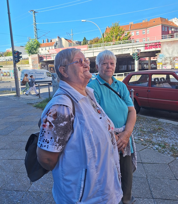 Zwei ältere Frauen mit kurzen Haaren und lockerer heller Kleidung stehen auf der Straße und freuen sich