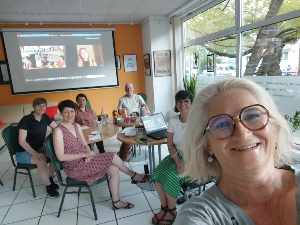 Im Vordergrund eine blonde Frau mit Brille, die das selfie macht, dazwischen Frauen auf Stühlen und im Hintergrund eine Videoleinwand mit weiteren Teilnehmenden