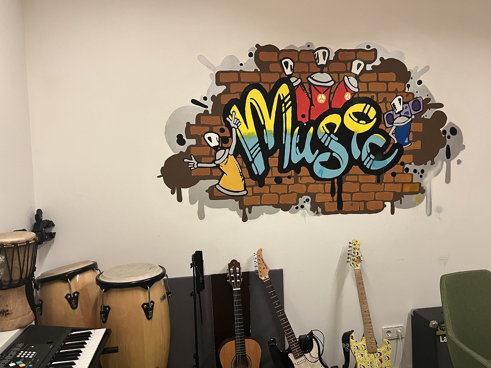 Im Zentrum des Fotos ist ein lustiges Wandgemälde mit dem Schriftzug Music zu sehen. Auf dem Fußboden darunter stehen verschiedene Musikinstrumente. Sie deuten darauf hin, dass wohl Musik gemacht wird in diesem Raum.