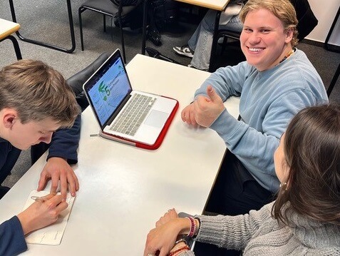Drei junge Menschen sitzen um einen Tisch mit einem geöffneten Laptop. Einer blickt in die Kamera und zeigt ein "Daumen hoch".