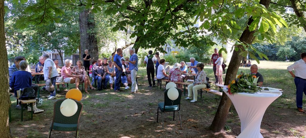 Menschen sitzen und stehen auf einer Wiese unter Bäumen an Biertischen und unterhalten sich.