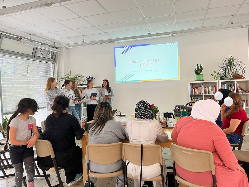 Die fünf Workshop-Leiterinnen stehen in einem hellen Raum. Neben ihnen ist eine Seite ihres Vortrags an die Wand projiziert. Darauf steht „Selbstverteidigung für Frauen“ sowie die fünf Namen. Vor ihnen sitzen mehrere Frauen und Mädchen und sehen sich den Vortrag an.