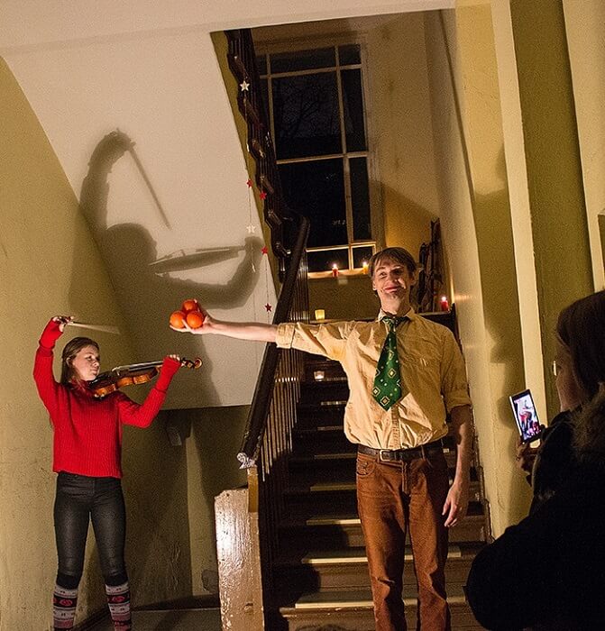 Impression einer musikalischen Aufführung im begehbaren Adventskalender: in einem Treppenhaus spielt eine junge Frau Violine, während ein Mann drei Apfelsinen in die Höhe hält.