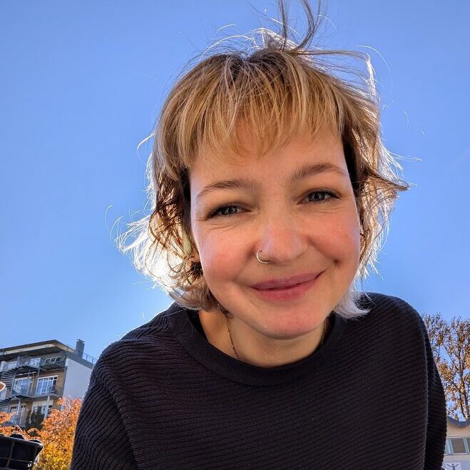 Jacqueline Kappl schaut lächelnd in die Kamera und beugt sich dabei leicht zum Betrachter nach vorne. Sie hat mittellange blonde Haare und trägt einen schwarzen Pullover.