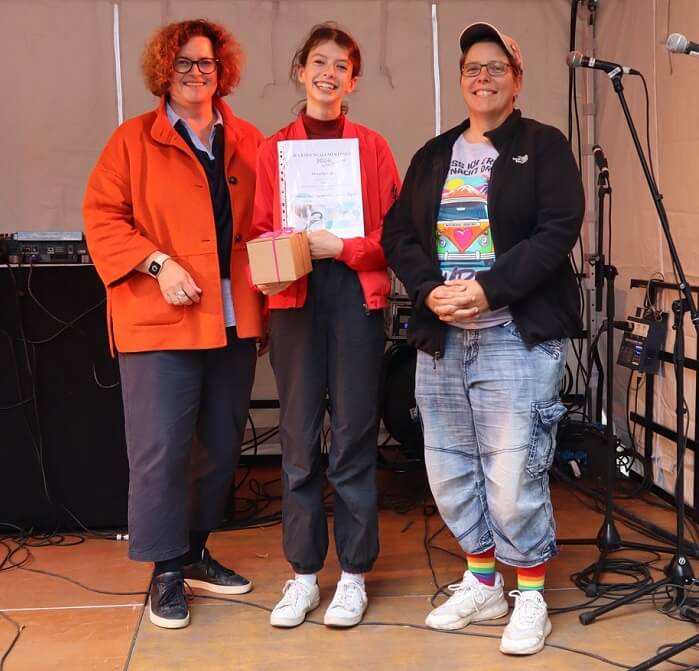 Die Preisträgerin Flora Spindler steht auf einer Bühne und zeigt lächelnd ihre Urkunde in die Kamera. Links und rechts neben ihr stehen die beiden Politikerinnen, die die Urkunde überreicht haben.
