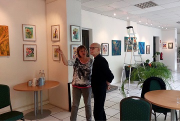 Die Künstlerin Ramona Emmerich und ein Mitglied des oskar Galerie-Teams stehen in der Ausstellung und unterhalten sich. An den Wänden hängen Bilder und im Hintergrund ist noch die Leiter zu sehen, die zum Aufhängen benötigt wird.