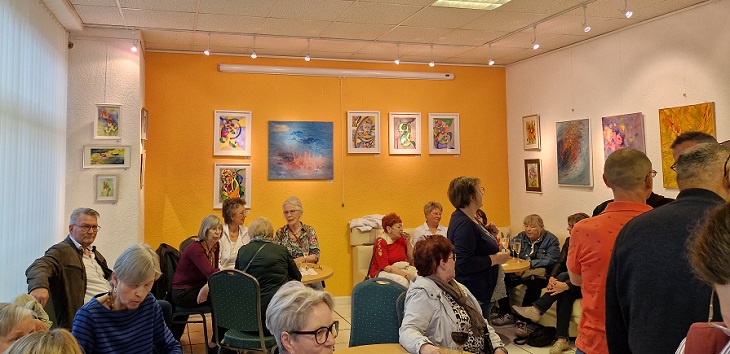 Viele Menschen stehen und sitzen in einem Raum, an dessen Wänden Kunstwerke hängen.