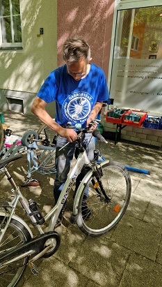 Ein Mann im blauen T-Shirt schraubt am Lenker eines Fahrrads. Im Hintergrund sieht man Werkzeug und ein umgedrehtes Fahrrad.