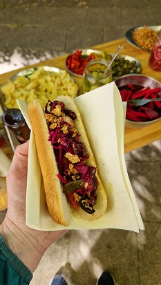 Jemand hält einen Hot Dog in der Hand. Im länglichen Brötchen sieht man Rotkohl, Zwiebeln und Champignons. Im Hintergrund ist ein Tisch zu sehen, auf dem die Zutaten zur Selbstbedienung aufgereiht sind.