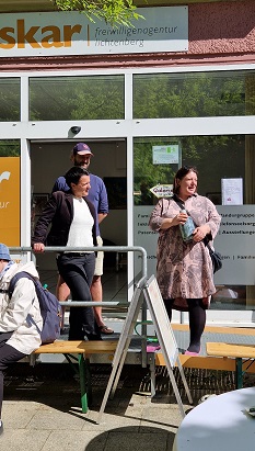 reden-oskar-nachbarschaftsfest-2025 Petra Grampe von der Stiftung Stadtkultur hält eine Rede und steht dabei vor dem Eingang der oskar | freiwilligenagentur in der Sonne. In den Händen hält sie eine Vase.