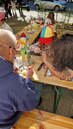 Ein grauhaariger Mann und ein Mädchen mit bunter Schirmmütze sitzen in der Sonne an einem Tisch und basteln Schmuck. Eine langhaarige Frau beugt sich zu dem Mädchen und hilft ihr. Auf dem Tisch liegen Fäden, bunte Perlen und Werkzeug.