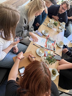 Mehrere Menschen sitzen um einen Tisch mit Kaffeetassen herum und unterhalten sich. Eine Person schreibt dabei etwas auf ein Blatt Papier.