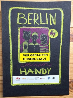 Auf schwarzem Hintergrund ist mit gelben und lila Wachmalstiften ein Handybildschirm gemalt. Darauf steht: "Berlin in meinem Handy" und "Wir gestalten unsere Stadt". Außerdem ein gemaltes Bild einer Familie.