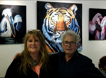 Die Künstlerin Silke Rossius mit Axel vom Galerie-Team vor einer Wand mit den Tiergemälden von Silke (Pelikan, Tiger und Flamingo).