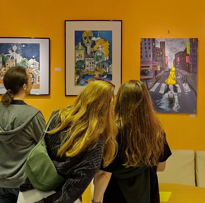 Besucherinnen einer Ausstellung schauen sich Bilder an einer orangefarbenen Wand an und stecken dabei die Köpfe zusammen.