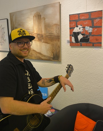 Der Sänger LordFerckel steht mit seiner Gitarre vor seinem Bild "Klaus der Panda" und lächelt in die Kamera.