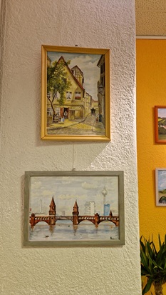 An der Wand hängen zwei Gemälde. Das obere zeigt eine Straßenszene mit historischem Haus. Das untere Bild zeigt die Oberbaumbrücke in Berlin.