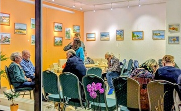 Blick durch ein großes Fenster in einen beleuchteten Raum an dessen Wänden Bilder hängen. Einige Personen sitzen auf Stühlen und unterhalten sich.