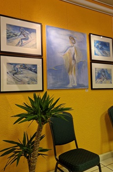 An einer hellorange gestrichenen Wand hängen fünf GEmälde. Das mittige ist am größten und zeigt vor blauem Hintergrund einen wahrscheinlich weibliche Person von hinten, die nur mit einem Tuch bekleidet ist. Die anderen vier Bilder sind links und rechts aufgehängt und zweigen jeweils einen Skifahrer bei der Abfahrt. Auch hier dominieren Blautöne.