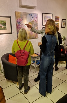 Drei Frauen stehen vor einer Wand mit Gemälden und Aquarellen. Sie sehen die Bilder an gestikulieren.