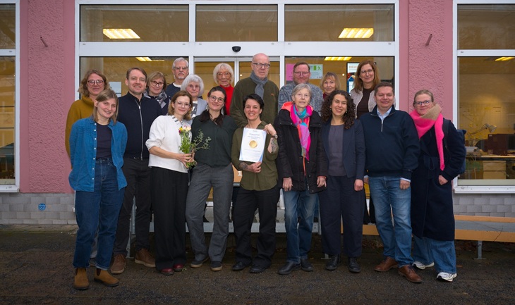 Eine Gruppe von Menschen steht in zwei Reihen vor einem Gebäude. Die Person vorne in der Mitte hält ein Blatt mit Logo des bagfa-Qualitätssiegels in Händen. Alle lächeln in die Kamera.