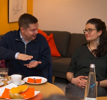 Ein Mann und eine Frau sitzen nebeneinander an einem Tisch und der Mann spricht und gestikuliert dabei.
