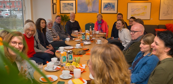 An einem langen Tisch sitzen zu beiden Seiten Menschen, die meisten lachen. Auf dem Tisch stehen Kaffeetassen, Teller, belegte Brote und Kuchen.