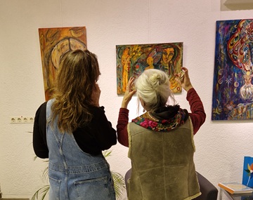 Anna Maria Michalska gestikuliert vor einem ihrer Gemälde und eine andere Frau schaut ebenfalls auf das Gemälde. Beide sind im Gespräch.