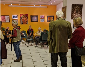 Blick in die Ausstellung. Zu sehen sind zwei Wände mit Gemälden und Zeichnungen. Im Raum stehen und sitzen Besuchende der Ausstellungseröffnung.