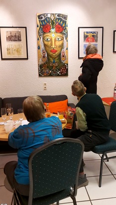 Drei Personen schauen auf ein Gemäöde an der Wand. Zwei von ihnen sitzen an einem Tisch und die dritte Person steht. Auf dem Gemälde ist in bunten Farben ein weibliches Gesicht mit Kopfschmuck zu sehen.