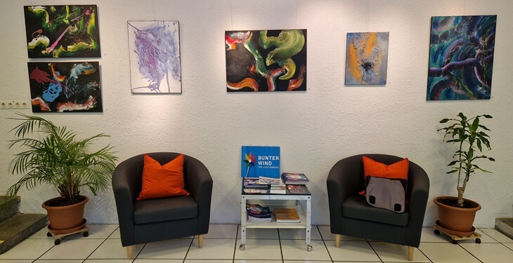 Blick in die Ausstellung "Energie trifft Resonanz" An der Wand hängen sechs abstrakte Bilder von Anne Leinen. Darunter stehen zwei Sessel neben einem Tisch mit Infomaterial.