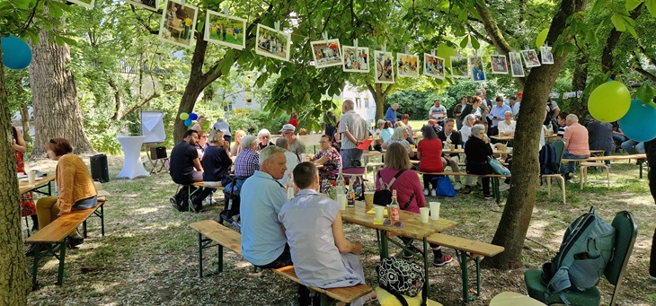 Menschen sitzen an Biertischen unter Bäumen und unterhalten sich.