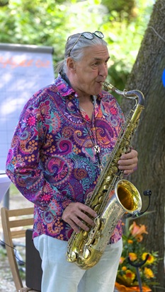 Ein Mann in buntem Hemd spielt Saxophon unter Bäumen.