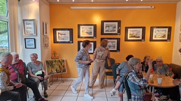 Einige Personen sitzen und stehen, unterhalten sich oder schauen Bilder an der Wand an.