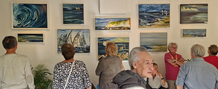 Besucher der Vernissage sehen sich Landschaftsgemälde an, welche in zwei Reihen an der Wand hängen.