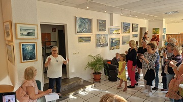 Ulrike Drobniewski steht vor einer Wand mit ihren Bildern und spricht zu den Gästen der Vernissage.