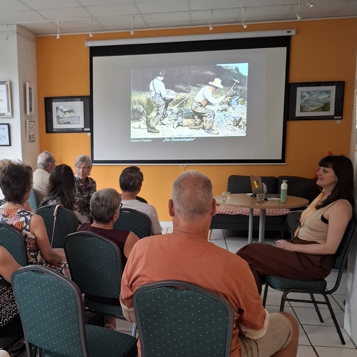 Besucher des Kunstplauschs sitzen auf Stühlen und schauen auf die Leinwand mit einem Kunstwerk.