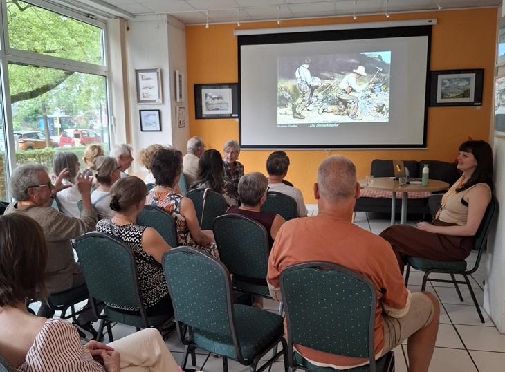 Besucher des Kunstplauschs sitzen auf Stühlen und schauen auf die Leinwand mit einem Kunstwerk.