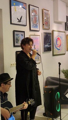 Eine Sängerin mit Mikrofon steht vor Kunstwerken, die an der Wand hängen. Neben ihr sitzt ein Gitarrenspieler.