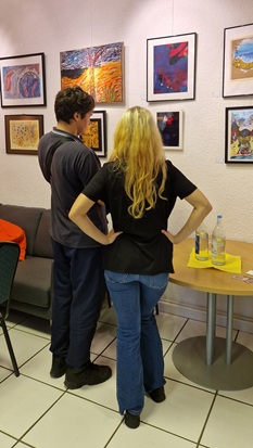 Zwei junge Menschen stehen vor einer Wand voller Kunstwerke und blicken darauf.